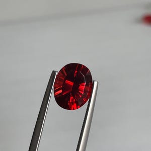 May include: A faceted, oval-cut, deep red gemstone is held by silver tweezers. The gem's internal structure reflects light, creating a brilliant display of color. The background is a soft, neutral gray.