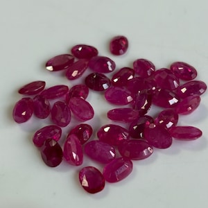 Puede incluir: Un conjunto de rubíes rojos profundos de corte ovalado. Las piedras preciosas están dispersas sobre una superficie blanca, cada una con facetas que capturan la luz. Los rubíes varían ligeramente en tamaño y tono.