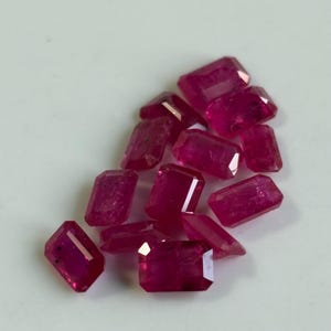 May include: A collection of faceted, rectangular-cut ruby gemstones. The rubies are a deep, vibrant red color and appear to be of varying sizes. The gemstones are scattered on a light-colored surface, showcasing their clarity and brilliance.