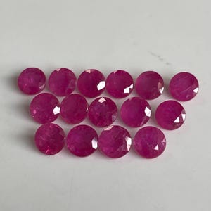 Könnte beinhalten: Eine Ansammlung von vierzehn runden, facettierten, leuchtend pink-roten Edelsteinen, die in drei Reihen angeordnet sind. Jeder Stein weist einen Brillantschliff mit zahlreichen dreieckigen Facetten auf, die das Licht reflektieren.