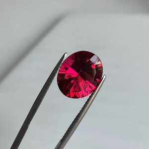 May include: A faceted, oval-shaped, deep red gemstone is held by a pair of silver tweezers. The gem has multiple facets that reflect light, creating a brilliant display of color. The background is a soft, neutral gray.