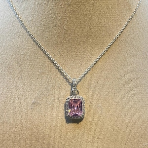 May include: A silver necklace with a rectangular pink gemstone pendant. The pendant is surrounded by a halo of small, clear stones. The chain is delicate and hangs on a neutral background.