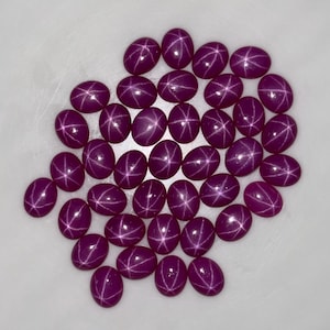 May include: A cluster of oval-cut, deep magenta gemstones, each displaying a star-like pattern. The gems are closely arranged, highlighting their polished surfaces and the star effect within. The background is a plain white surface.