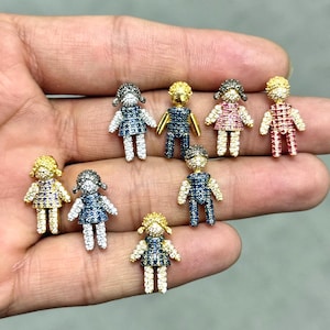 Puede incluir: Una colección de pequeños colgantes con forma de muñeca. Los colgantes están adornados con piedras preciosas brillantes en varios colores, incluyendo oro, plata, azul, rosa y rojo. Cada muñeca tiene un vestido y peinado detallados, creando un diseño encantador.
