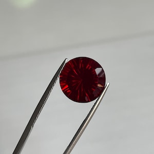 May include: A circular, faceted, deep red gemstone is held by silver tweezers. The gemstone features a radial pattern of cuts, creating a starburst effect. The background is a soft, neutral grey.