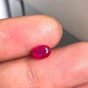 May include: A loose, oval-cut gemstone with a vibrant, deep red colour and faceted surface. The stone displays a polished finish with clear light reflection on its top edge, showcasing a translucent quality and a saturated, uniform crimson hue.