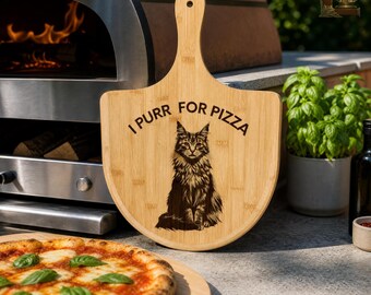 May include: A light brown wooden pizza peel with a handle, featuring a cat illustration and the text "I Purr For Pizza." The peel is near a pizza oven and a freshly baked pizza.