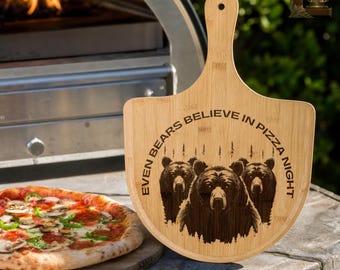 May include: A wooden pizza peel with a handle, featuring the engraved text "EVEN BEARS BELIEVE IN PIZZA NIGHT" and a graphic of three bears. A pizza with toppings sits on a wooden board.