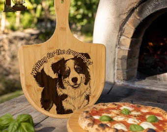 May include: A wooden pizza serving board with a handle, engraved with a dog image and the text "Waiting for the droppings!". A pizza with tomato sauce, mozzarella, and basil is on a wooden board. The scene is set outdoors near a pizza oven.