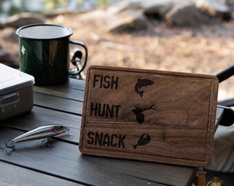 May include: A rectangular wooden chopping board with the words "FISH", "HUNT", and "SNACK" etched into the surface. The board also features etched images of a fish, deer, and a piece of meat. A fishing lure and a green speckled mug are also visible.