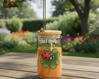 May include: A glass jar with a bamboo lid and a clear straw, filled with an orange liquid. The jar features a four-leaf clover with a ladybug and the words "LIQUID LUCK". A small ladybug sits on the lid. The jar is on a wooden table.