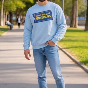 May include: A light blue sweatshirt with the text "Respect Your Elder Millennials" in a blue and yellow rectangle. The person is wearing light blue jeans and white sneakers. The person is standing outside on a sunny day.