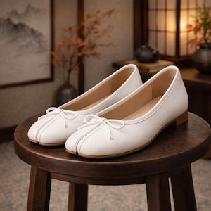 May include: A pair of white leather ballet flats with a split-toe design and small bow detail. The shoes have a low heel and are displayed on a wooden stool. The background features a painting and decorative elements.