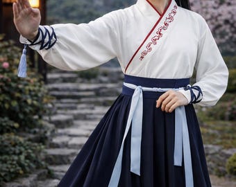 May include: A traditional white and navy blue outfit with long sleeves and a flowing skirt. The top has red trim and embroidered details. The outfit includes a navy blue sash with light blue ribbons and decorative accents on the sleeves. A light blue tassel hangs from the sleeve.