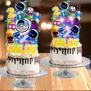 May include: Two round cakes with white frosting, chocolate ganache drips, and colorful sprinkles on glass stands. Each cake features a large, multi-character topper with the name CHLOE in yellow letters and the number 2, showcasing various robot figures.