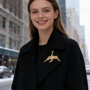 May include: A gold-colored bird brooch is pinned to a black coat. The brooch features a stylized bird with outstretched wings and a small, decorative element above its head. The background shows a snowy city street with buildings and pedestrians.