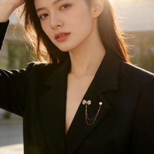 Könnte beinhalten: Ein schwarzer Blazer mit einer dekorativen Brosche. Die Brosche hat ein Schleifendesign mit einer kleinen Perle und herabhängenden Ketten. Der Blazer hat einen klassischen Schnitt und wird offen getragen. Das Bild wird von Sonnenlicht beleuchtet.