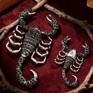 Könnte beinhalten: Zwei Skorpion-Broschen. Die größere ist schwarz mit schwarzen Edelsteinen und silberfarbenen Beinen und Klauen. Die kleinere Brosche hat einen silberfarbenen Körper und Beine mit schwarzen Edelsteinen. Beide liegen auf rotem Samt.