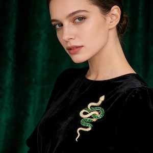 Könnte beinhalten: Goldfarbene Schlangenbrosche mit smaragdgrünen und klaren Kristallakzenten. Das Schlangendesign ist aufwendig detailliert, mit einem gewundenen Körper und einem erhobenen Kopf. Die Brosche ist vor einem schwarzen Samthintergrund platziert.