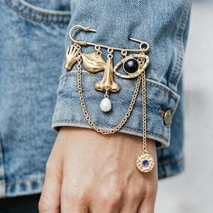 Könnte beinhalten: Eine goldfarbene Brosche mit Sicherheitsnadel-Design und hängenden Anhängern. Zu den Anhängern gehören eine Hand, Lippen, eine Nase und ein Auge mit schwarzem Zentrum. Eine Kette mit einem blauen Edelsteinanhänger hängt darunter. Die Brosche ist an einer Jeansjacke befestigt.