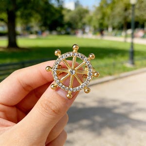 Könnte beinhalten: Goldfarbene Schiffsrad-Brosche mit klaren Kristallakzenten. Die Brosche hat ein kreisförmiges Design mit Speichen und einer zentralen Nabe und wird in einer Hand vor einem verschwommenen Hintergrund gehalten.