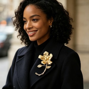 Könnte beinhalten: Eine goldfarbene Blumenbrosche mit Perlenmitte und detaillierten Blättern. Die Brosche ist an einem schwarzen Mantel befestigt. Das Blumendesign ist ein klassisches Accessoire, das jedem Outfit einen Hauch von Eleganz verleiht.