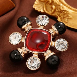 Könnte beinhalten: Goldfarbene Brosche mit einem quadratischen roten Mittelstein, umgeben von klaren Kristallen und schwarzen, kugelförmigen Akzenten. Die Brosche hat ein detailliertes, verziertes Design, passend für formelle Anlässe.