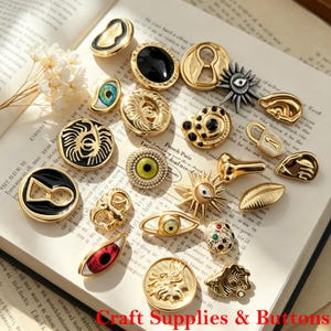 May include: A collection of assorted metal buttons featuring various designs including eyes, keyholes, sunbursts, and abstract shapes in gold, black, and silver tones. The buttons are arranged on an open book with printed text and white flowers.