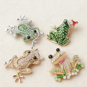 May include: Four frog brooches in various designs. One is silver with green and clear crystals, another is gold with clear crystals, and a third is green with a red hat. The fourth brooch is gold with green and clear crystals.