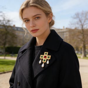 Puede incluir: Broche de cruz dorada con detalles rojos, verdes y blancos. El broche presenta un diseño de cruz con una gota de perla. El broche está prendido en un abrigo negro.