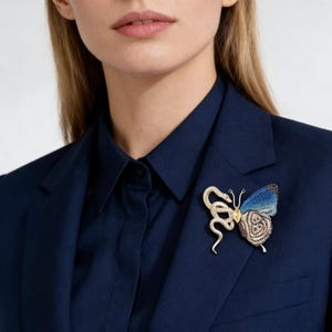 Può includere: Spilla dorata con un design a farfalla e serpente. La farfalla ha ali blu e marroni con un centro giallo. Un serpente dorato si avvolge attorno alla farfalla. La spilla è appuntata su un blazer blu navy.