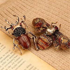 Puede incluir: Dos broches de insectos ornamentados. Uno es un escarabajo con cuerpo marrón, patas doradas y detalles negros y rojos. El otro es una abeja con un gran cristal y cristales ámbar y rojos. Ambos sobre una superficie texturizada.