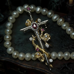 Könnte beinhalten: Eine detaillierte Brosche in Form eines Schwertes mit einem zentralen Rubin und Diamantakzenten. Das Schwertdesign ist mit goldfarbenen Rosen und weiteren Rubin-Details verziert. Eine Perlenkette umrahmt die Brosche.