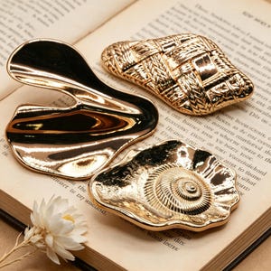 May include: Three gold-colored decorative objects rest on an open book. One is a curved, abstract shape, another is a woven rectangle, and the third is a seashell shape with a spiral design.