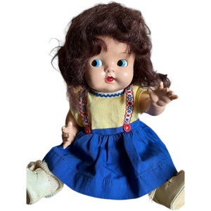 Puede incluir: Una muñeca vintage con cabello castaño, ojos azules y boca roja. Lleva una camisa amarilla con tirantes rojos y azules, una falda azul y zapatos blancos.