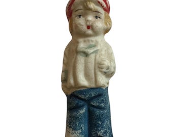Puede incluir: Una pequeña figurita de cerámica de un niño que lleva un sombrero rojo y blanco, una camisa blanca y pantalones azules. La figurita tiene una cara pintada con labios rojos.