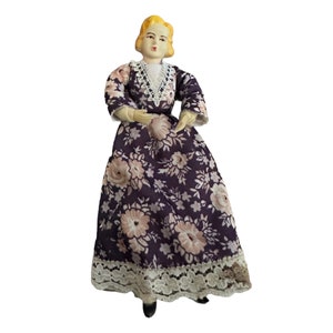 May include: A porcelain doll wearing a purple dress with a floral pattern and white lace trim. The doll has blonde hair and a painted face.