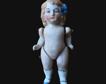May include: A porcelain doll with blonde hair, blue eyes, and a blue flower on her head. She is wearing white socks and blue shoes.