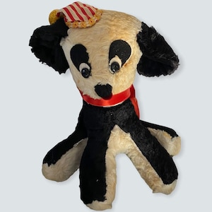 Puede incluir: Un perro de peluche vintage que lleva un sombrero a rayas rojas y blancas con una banda amarilla. El perro tiene pelo blanco y negro y grandes ojos negros.