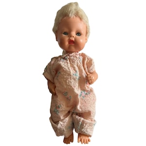 Puede incluir: Una muñeca de bebé de plástico vintage con cabello rubio, vestida con un pijama de estampado floral rosa y blanco. La muñeca tiene ojos azules y una sonrisa pintada.