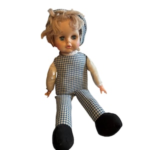 Puede incluir: Una muñeca vintage con cabello rubio, ojos azules y un atuendo de cuadros vichy. La muñeca lleva un vestido de cuadros vichy azul y blanco y un sombrero de cuadros vichy azul y blanco. La muñeca tiene zapatos negros.