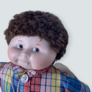 Pode incluir: Uma boneca de porcelana com uma peruca de cabelo marrom e felpudo, vestindo uma camisa xadrez com um colar de corrente dourada.