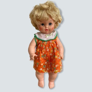 May include: A vintage doll with blonde curly hair wearing an orange dress with white and green floral print. The doll has blue eyes and rosy cheeks.