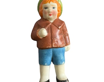 Puede incluir: Una figurita de porcelana de un niño que lleva una chaqueta marrón, pantalones azules y un sombrero verde. El niño tiene el pelo rojo y la cara pintada.