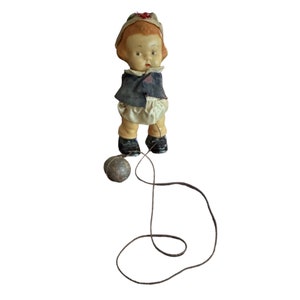 May include: A vintage pull toy with a porcelain doll dressed in a blue jacket and white pants. The doll is attached to a string that is connected to a weighted ball.