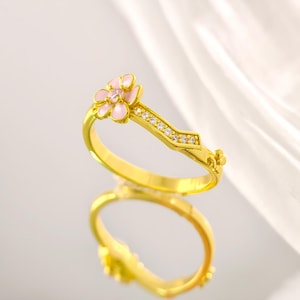 May include: A gold-colored ring featuring a pink and white flower design. The ring has a band adorned with small, clear gemstones. The flower has pink petals and a central gemstone. The ring is displayed on a reflective surface.