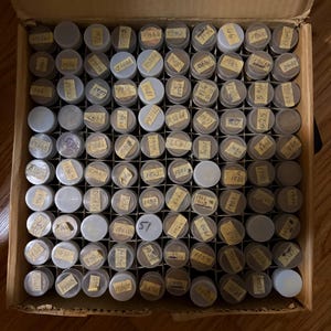 May include: A cardboard box filled with numerous small, clear plastic containers. Each container has a gray lid and a small, yellowed label with handwritten numbers. The box is open, revealing the contents, which appear to be collectible coins.