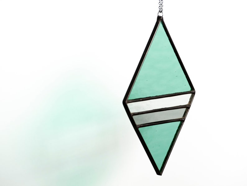 Geometric, modern stained glass suncatcher made with green, grey and clear wavy textured glass. Made in the Tiffany Glass style and with black patina. Hangs on a black chain on a white background to show color and texture of glass art. Side view.