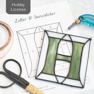 Può includere: Un acchiappasole a forma di lettera "H" in vetro colorato con vetro verde e bianco, circondato da una cornice nera. Sono visibili un modello e degli strumenti, tra cui forbici e un tagliavetro. L'immagine include il testo "Hobby License" e "Letter 'H' Suncatcher".