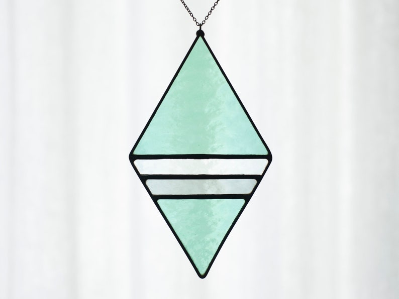 Geometric, modern stained glass suncatcher made with green, grey and clear wavy textured glass. Made in the Tiffany Glass style and with black patina. Displayed hanging from black chain in front of a white curtain.